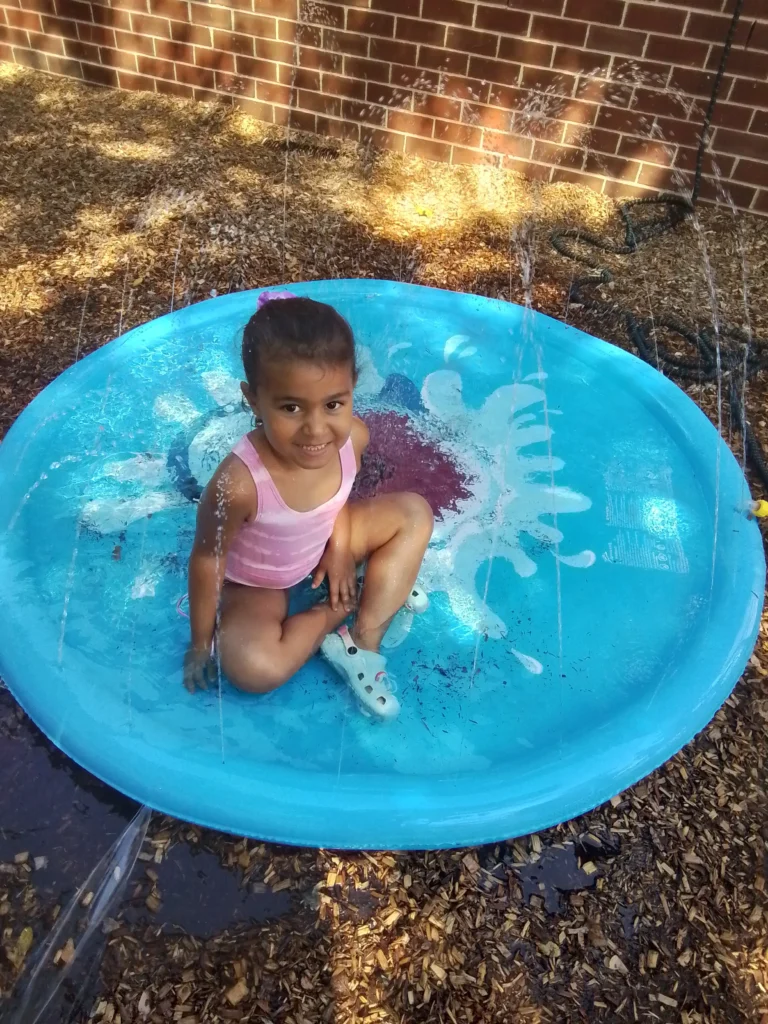 Young girl in a pink swimsuit sits in a bright blue round kiddie pool as water sprays from a sprinkler against a brick wall nearby.
