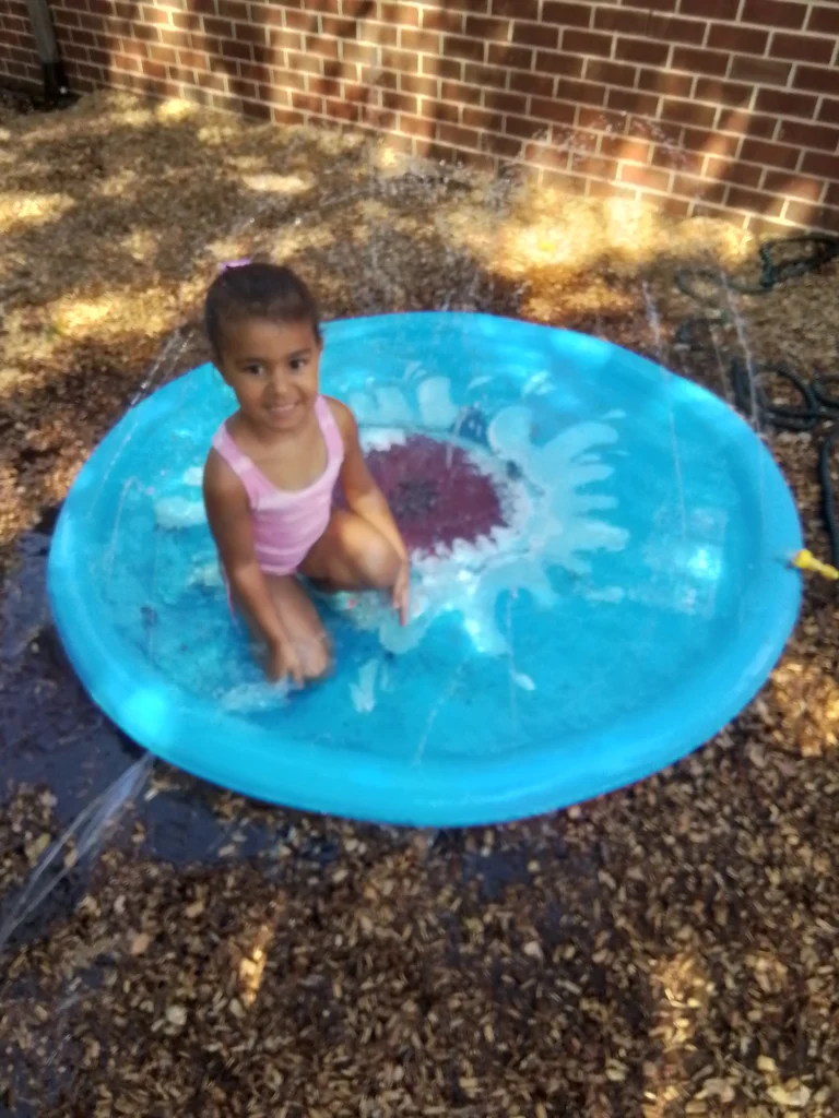 Young girl in a pink swimsuit sits in a blue plastic kiddie pool outdoors near a brick wall.
