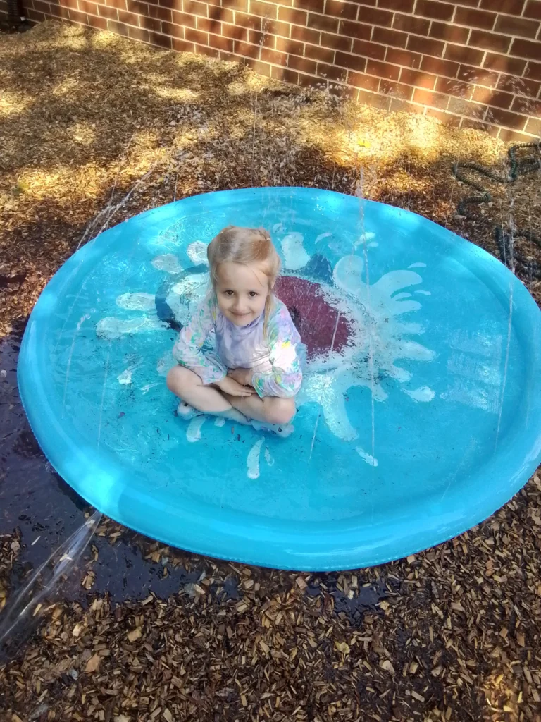 Young girl sitting cross-legged in a blue round water tub outdoors on wood chips by a brick wall, smiling at the camera.