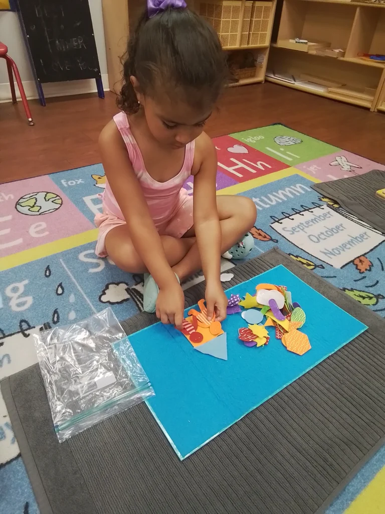 Young girl in a pink dress sits cross-legged on a colorful classroom rug, making a paper collage with orange shapes on a blue board.