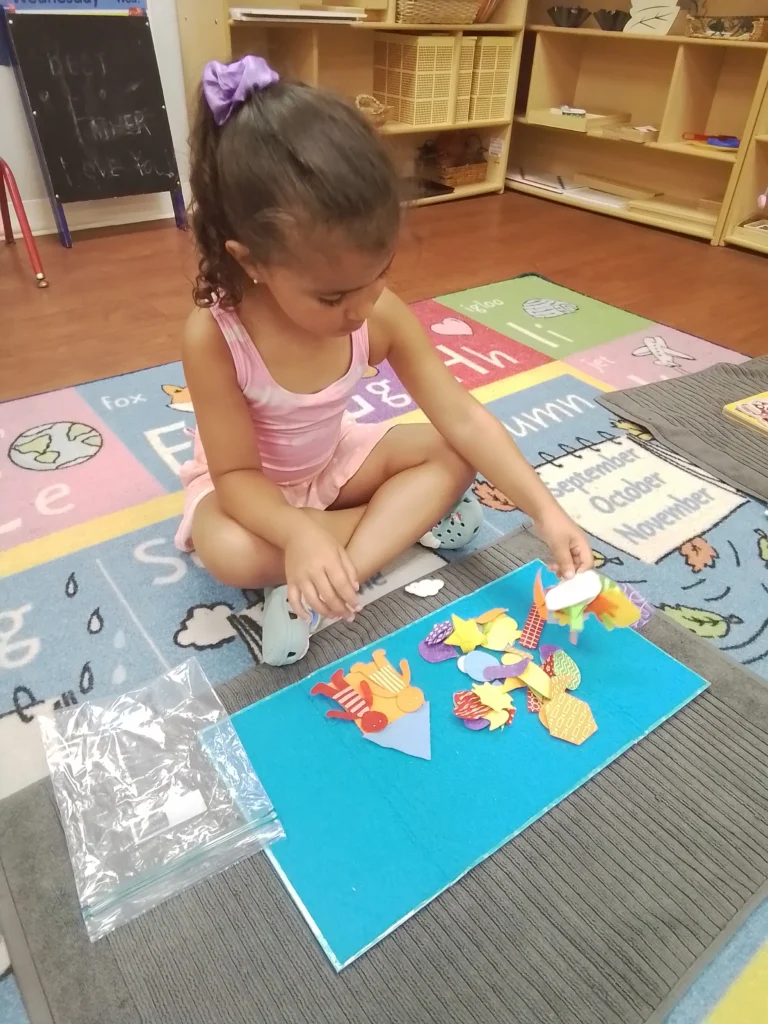 Young girl in a pink top sits on a colorful classroom rug, arranging paper shapes for a craft project.