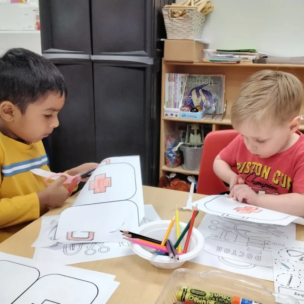 Two children sit at a table cutting out shapes from paper with scissors during a craft activity in a classroom.