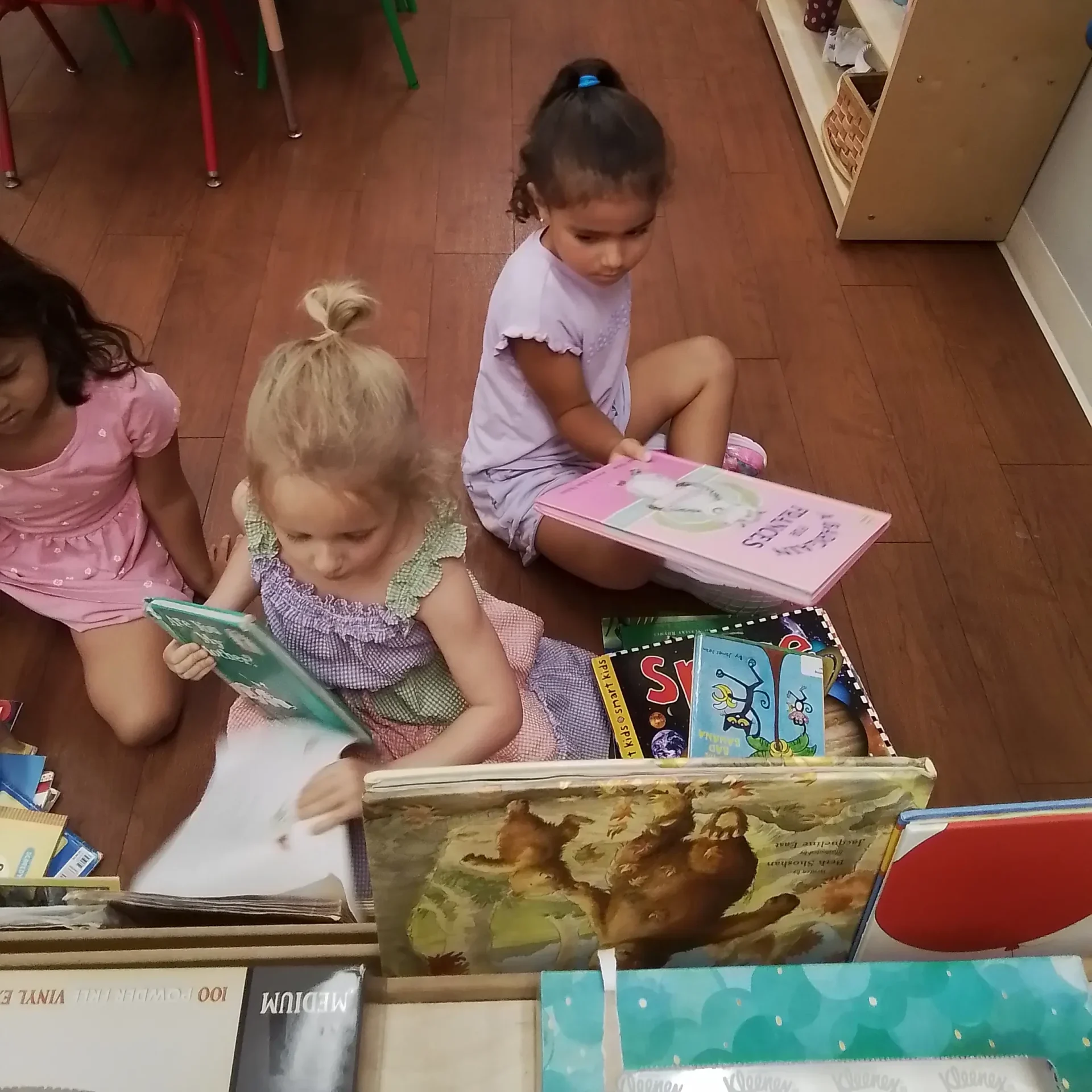 Children sit on a wooden classroom floor, reading colorful picture books; left: pastel tables and red/green chairs, right: shelves with water bottles and supplies.