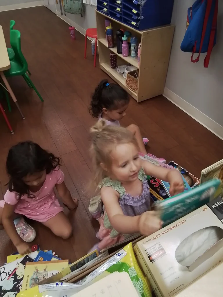Children sitting on a wooden classroom floor, looking through colorful books in a small reading area with shelves and backpacks nearby to the wall.