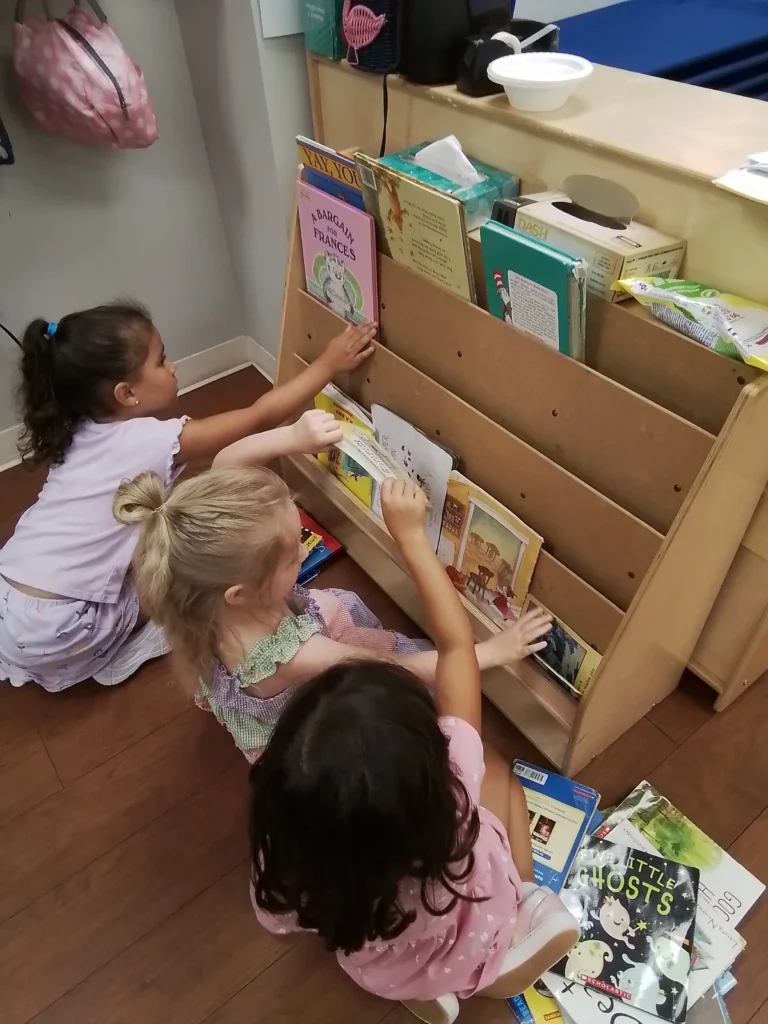 Children sitting on the floor by a wooden book display, browsing colorful picture books including a 'Little Ghosts' title visible.