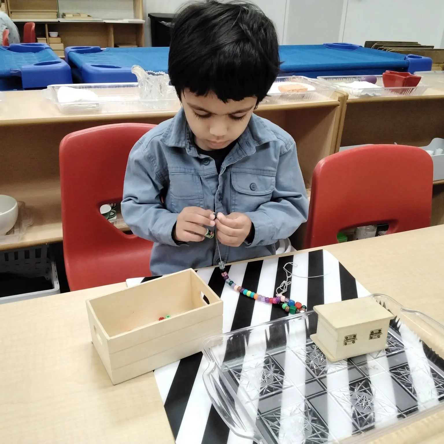 Young child in a denim shirt threads colorful beads to make a necklace at a classroom table.