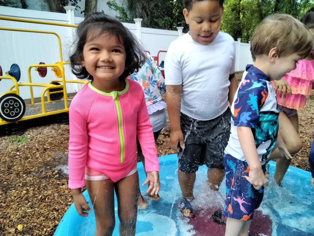 Group of children in swimsuits standing in a shallow splash pool outdoors, water splashing around near a yellow play structure and white fence.