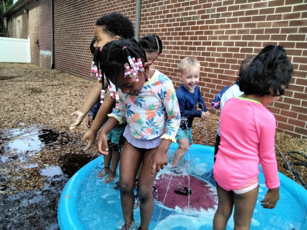 Group of children standing barefoot in a blue plastic tub filled with water, outdoors beside a brick wall, engaged in a water play activity.