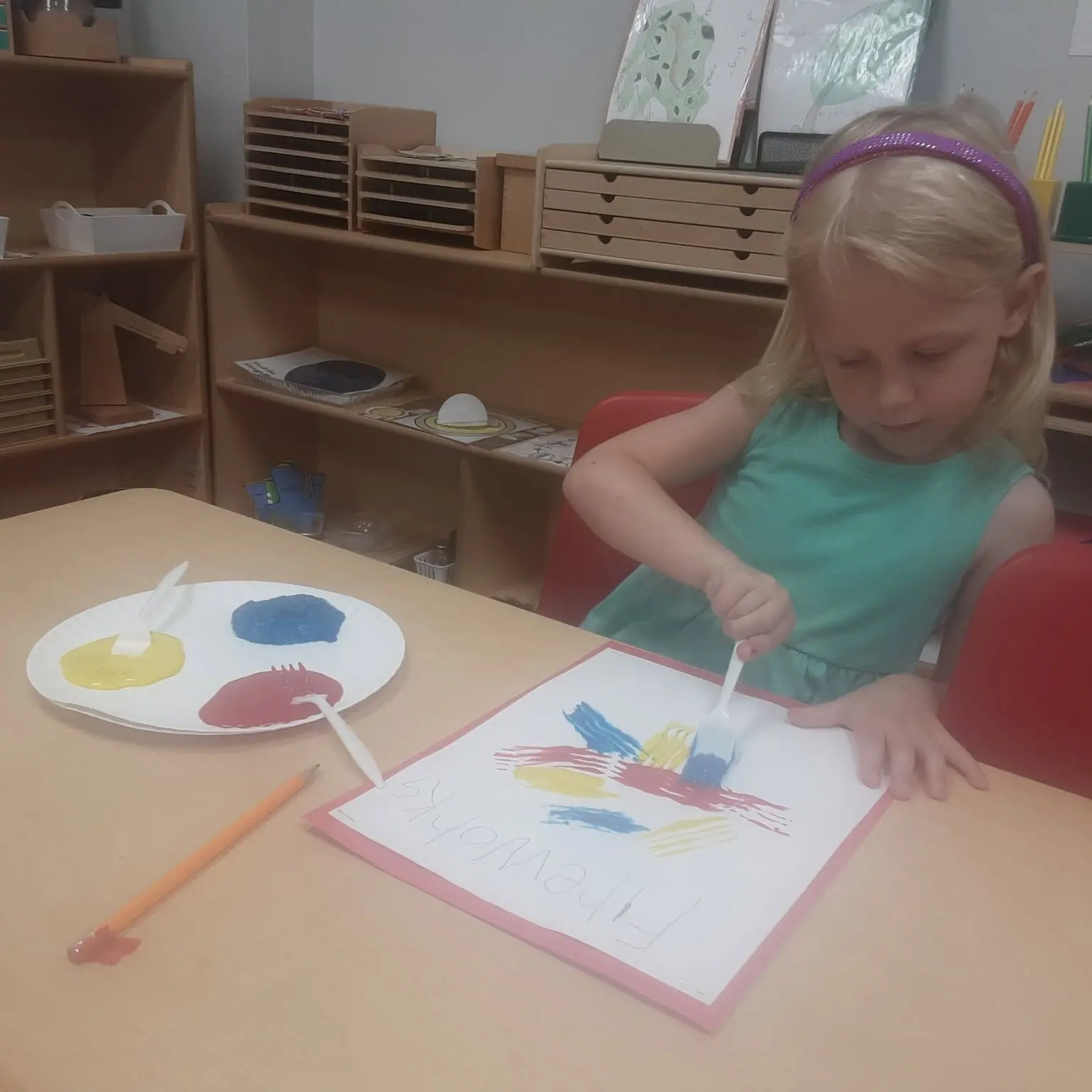 Young girl wearing a turquoise dress sits at a table painting with brushes; plate shows yellow, blue, and red paint nearby in a classroom.