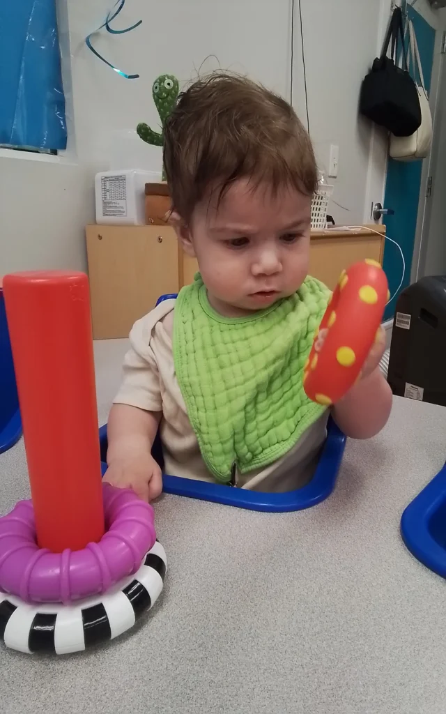 Baby seated in a high chair, wearing a green bib, holding a red toy with yellow polka dots and looking at it curiously in a play area.