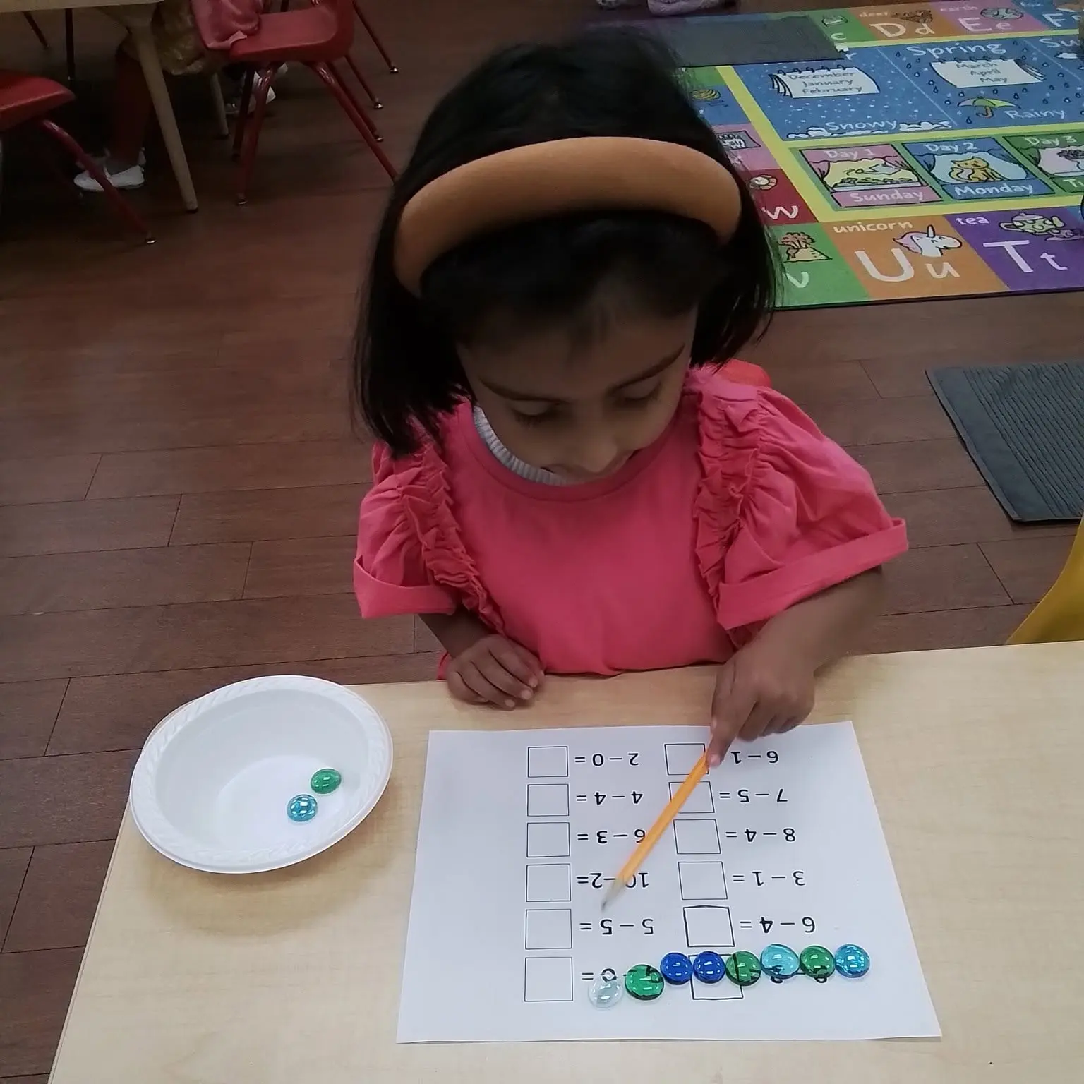 math-1 - Pinebrook Montessori Daycare and Preschool - Pinebrook Montessori Daycare and Preschool Young girl in a pink top sits at a table, solving a math worksheet with a pencil, while colorful marbles are arranged in a row nearby in a white bowl and on the paper.