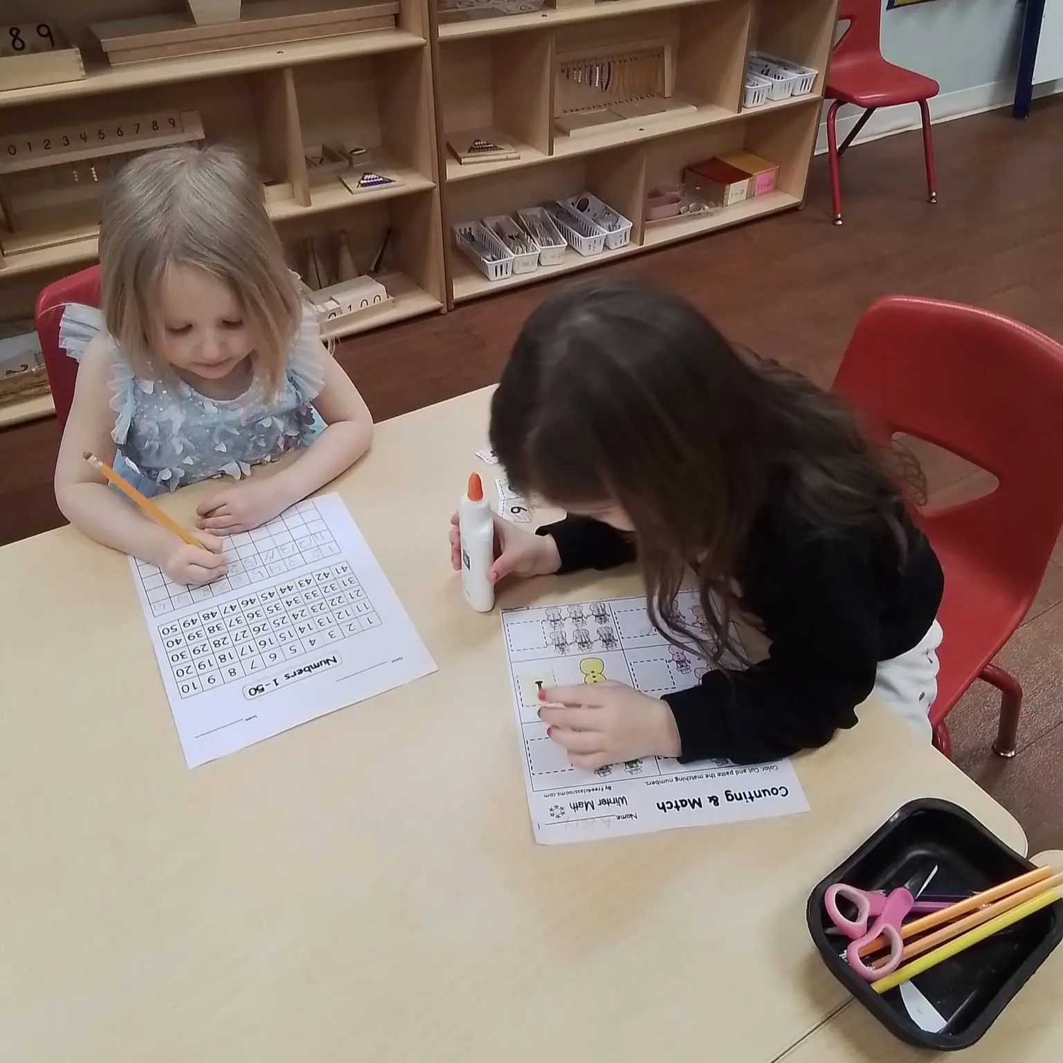 Two young girls sit at a classroom table, each working on worksheets with pencils and glue sticks, shelves of educational toys in the background.