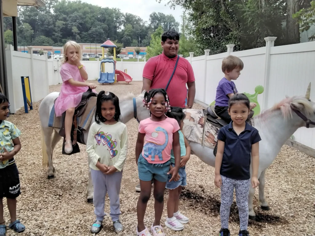 Group of diverse children riding ponies together in a outdoor play area, with a man supervising in a red shirt and playground equipment in the background.