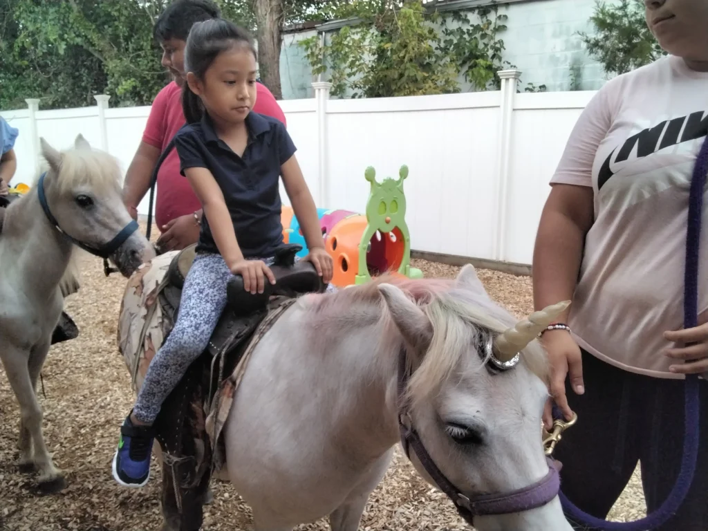 Young girl in a navy polo shirt riding a gray pony as adults stand nearby in a fenced backyard.