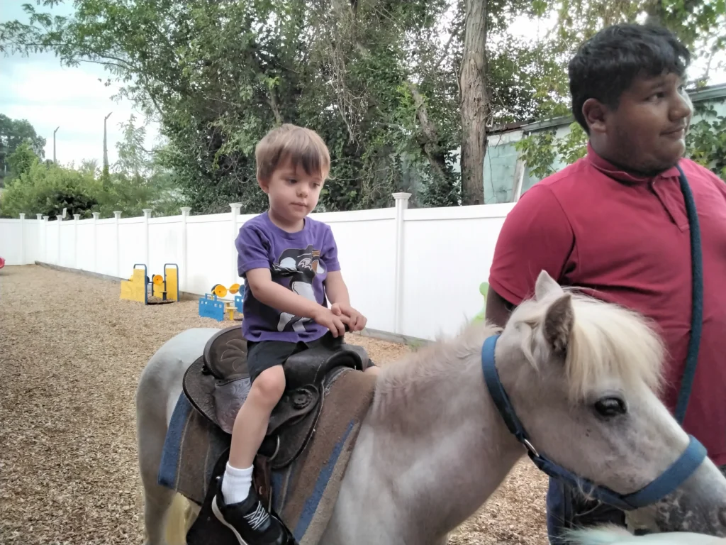 A young boy in a purple shirt rides a light gray pony, guided by a man in a red shirt outdoors near a white fence and trees.