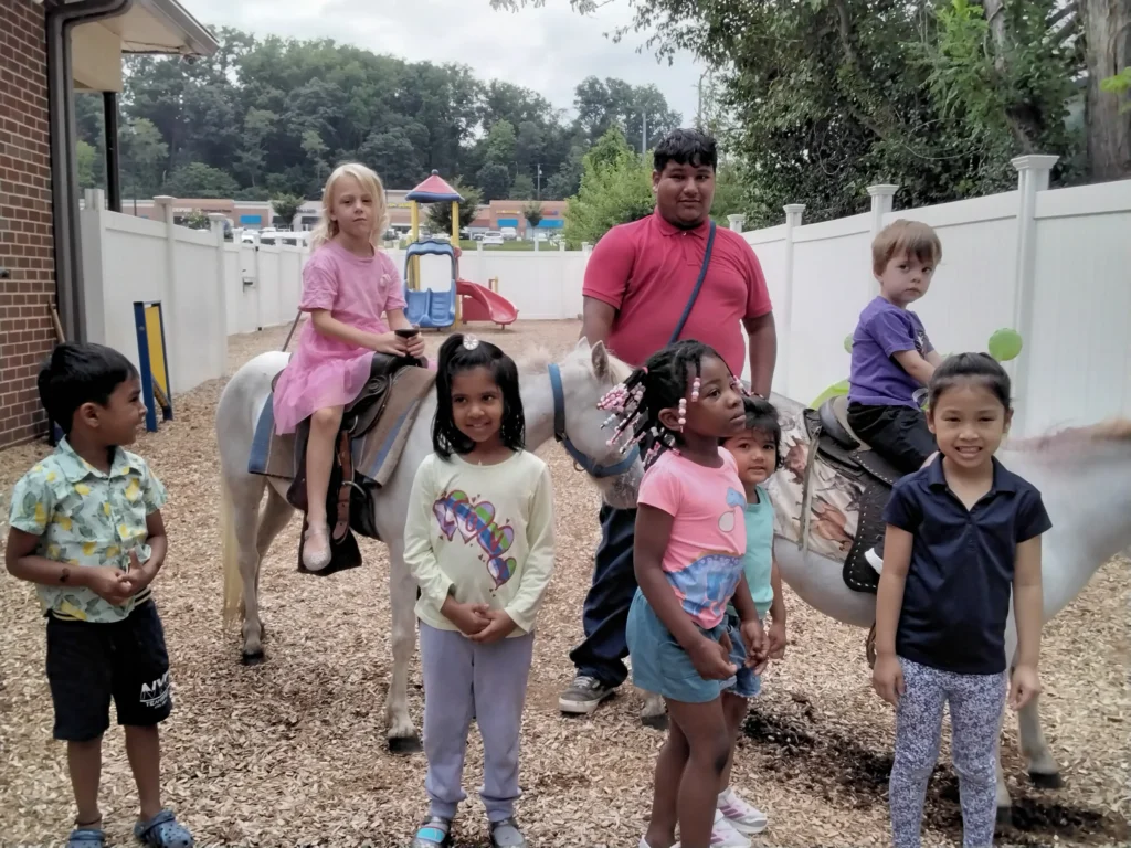 Group of children riding horses in a fenced playground area, with a man in a pink shirt standing beside them.