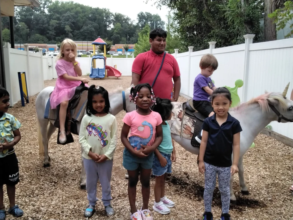 Group of children riding ponies in a fenced outdoor play area, with a man supervising nearby.