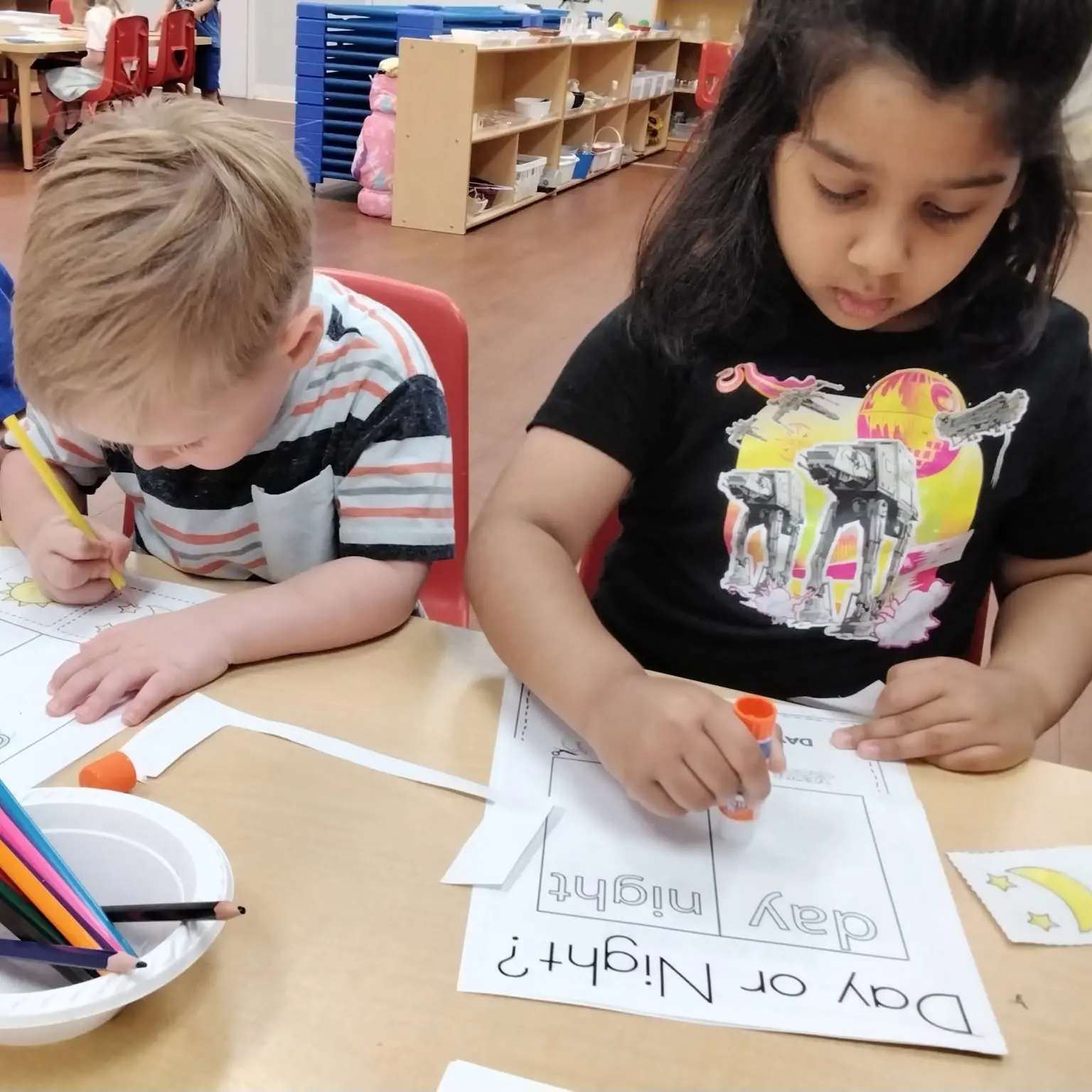 pre-k-1 - Pinebrook Montessori Daycare and Preschool Two children sit at a table coloring worksheets in a classroom, using pencils and glue sticks.