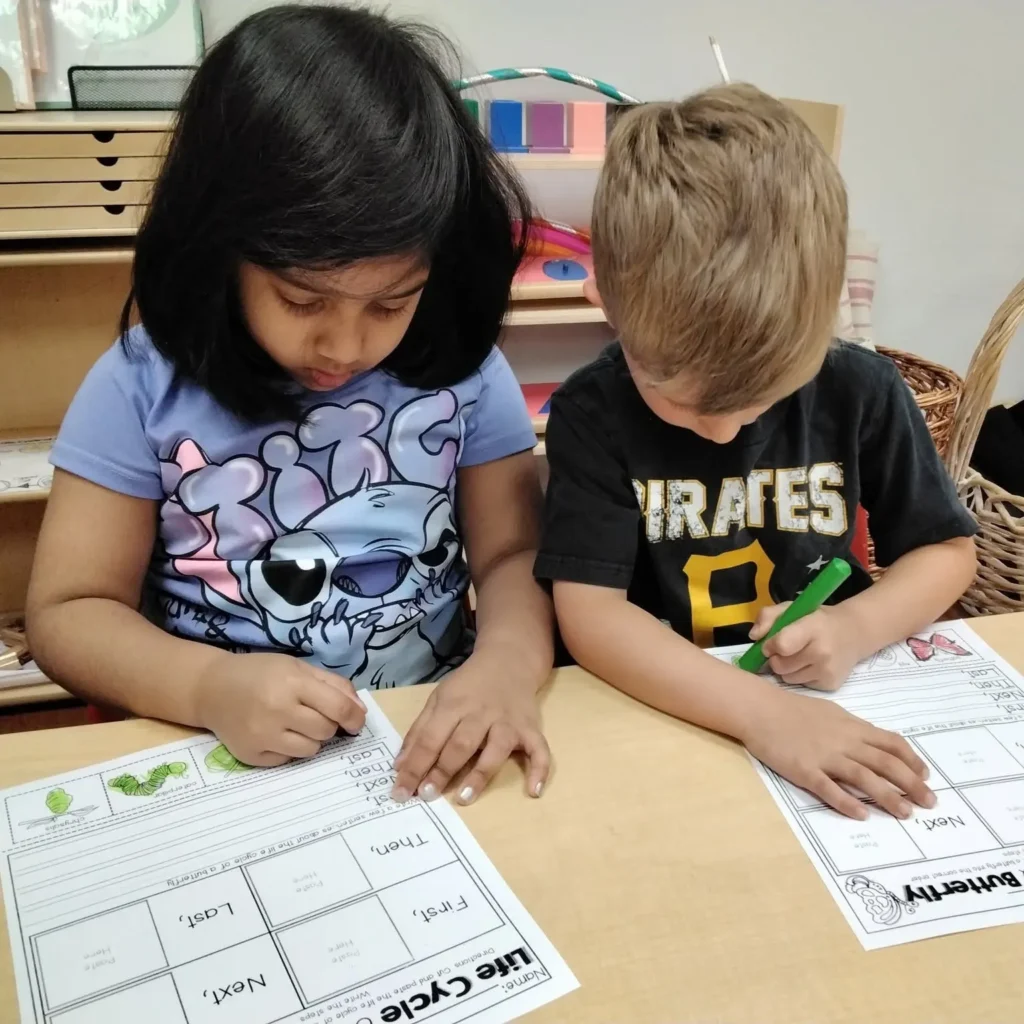 Two children sit at a wooden table, writing and coloring on alphabet worksheets during a learning activity at school or home.