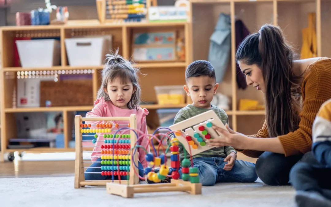 Teacher watches two preschoolers playing with a colorful bead maze in a classroom setting.