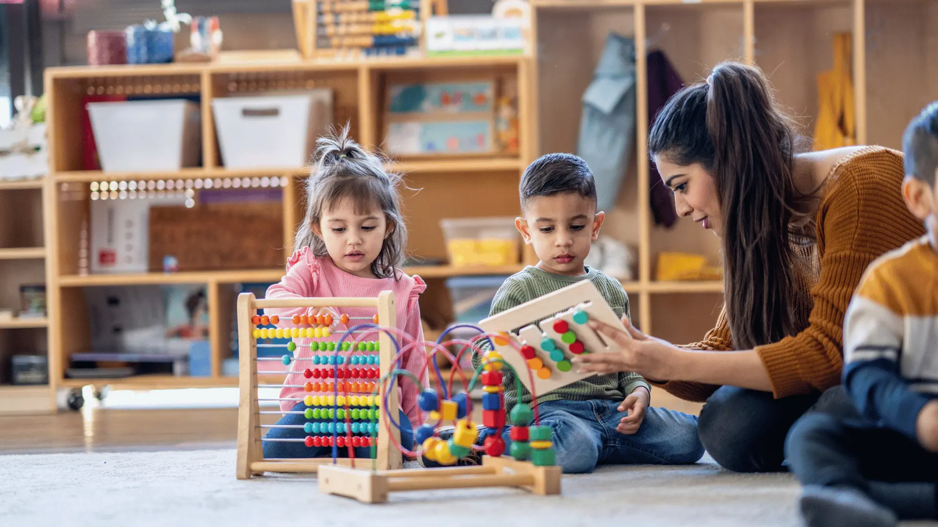Teacher watches two preschoolers playing with a colorful bead maze in a classroom setting.